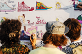 A color photo of three young New Yorkers stand before large piece of white poster paper covered with images of Converse All-Star hightop shoes, which they are applying their own designs too with different colored markers.
