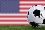 A soccer ball on grass in front of the U.S. flag