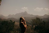 Woman with her hands by her chin, contemplative. She’s in the center of the photo, side profile. There are mountains behind her.