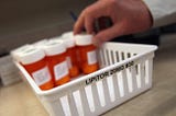 A hand is seen reaching for a white plastic basket containing multiple orange prescription pill bottles. A label on one of the bottles prominently reads “LIPITOR 20MG #30”. The setting appears to be a pharmacy or medical facility.