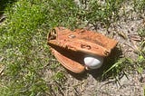 Photo of an old Louisville Slugger baseball glove lying on the ground which is a mixture of dirt and grass. The glove has a white baseball in it that is halfway visible.