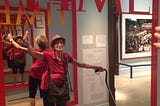 Woman in a hat with a can with her arms spread out in front of a large mirror that says “Time” (as in the magazine). Museum setting.