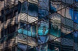 “Blue and white concrete building”, photograph by Robert Ullman (source: https://artist.scop.io/image/blue-and-white-concrete-building-73) — a building façade of square glass panes reflecting a distorted image of a concrete building in shades of dark blue, aqua, and off-white.