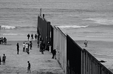 Black-and-white photograph of people on a beach, divided by a tall fence.