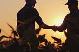 Two farmers stand in a field and shake hands in silhouette at sunset.