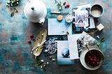 An aerial view of a writing desk, with a pen, a feather, several holiday cards, a teapot and cup of chocolate and a bowl of berries all arranged together.