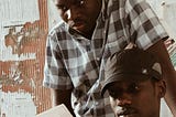 man in black and white plaid shirt peers into a a copy of The Punch newspaper held by another man in black face cap and black and white plaid shirt