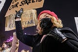 Several women protesters standing in the street, one blonde woman with a black mask holding a sign saying, “Our Bodies, Our Streets.”