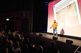 Gordon Fong standing on stage with a microphone in hand explaining about the tweet behind him on a big screen.