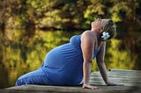 Color photo of a pregnant woman in a royal blue sleeveless dress with a flower in her hair. She’s relaxing on the edge of a boat dock, in the summer, enjoyinh  the moment.