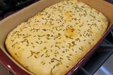 Image of baked focaccia, topped with sea salt and fresh rosemary, in stoneware pan