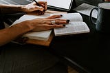 A woman’s hand holding a book open and writing in a journal.