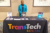 E.C. Pizarro III, a Black man wearing a teal jacket, smiles while standing behind a table with fliers, buttons, and a black drape reading “TransTech Social” in rainbow-colored lettering.