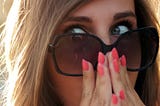 Blonde woman has a surprised face. She is looking over her big sunglasses and she is holding both hands (with painted fingernails) in front of her face.