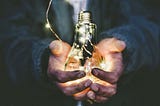 a person with dirty hands holds a glowing lightbulb against a dark background
