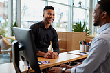 Two colleagues at a desk — one smiling and making eye contact with a customer while the other types into a computer system.
