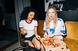 Two women eating pizza. They look like they’re having fun together. The image represents the idea that we give data away in everyday life without thinking. Free pizza, with a side of cyber vulnerability.