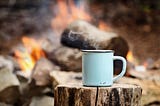 An enamel mug containing a hot drink sitting on top of a log.
