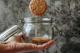 Hand holding cookies jar with one cookie inside and one falling into the jar.