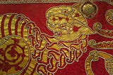 Detail of an embroidered mantle. The fabris is dyed deep red and gold embroidery depicts a stylised lion with a mane that looks like flames and a striped body. The finer details are picked out in double rows of pearls.