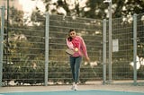 A female in yoga pants and a red sweatshirt returns a pickleball on a pickleball court.