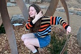 Nona is balancing on a metal abstract sculpture in a crouched position. Her left hand is holding onto the sculpture and her right is brushing her hair out of her face. She is laughing looking away at the camera and wearing a striped sweater and a jean skirt.