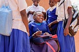 A young girl in a blue sweater sits in a wheelchair smiling surrounded by other school friends in white and blue uniforms all smiling