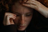 a sad woman gazing downward with one hand touching her forehead and the other wiping her eye