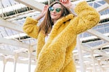 A woman wearing a yellow fake fur coat, green lensed sunglasses, and feeling herself.