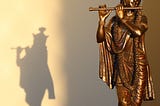 A full length Krishna statue is placed to the right of the image with sunlight falling on it in an angle from the top making it shine and there is a shadow of the statue falling to the left of the image.