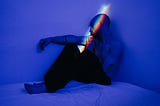 A man sits on top of a bed in a room lit with blue light. A rainbow projection is lighting up his face.