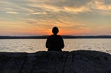 The author is sitting by a lake, facing the sunset in Sweden