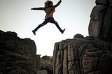 A person jumping from a large boulder to another large boulder over a sudden drop.