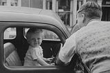 Man looking into an old car with a small boy at the driving seat looking at the camera smiling. Black and white photo.