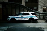 A Chicago Police SUV parked along a city street at night outside a stone and glass building, its blue-and-white markings and “Chicago Police” lettering clearly visible under dim street lighting.