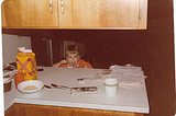 The author as a boy, eating a bowl of oatmeal. The boy wears an orange shirt and there is a yellow paper milk carton to the his right, the viewer’s left