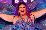 A screenshot of a smiling Spankie Jackzon with her arms outstretched from season 2 of RuPaul’s Drag Race Down Under, wearing a purple, blue and pink handcrafted costume that somewhat resembles a butterfly.