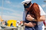 A heartfelt reunion unfolds as a person in a U.S. Navy uniform shares a tender hug with a woman with fiery red hair, clad in a polka-dot dress, against the backdrop of a bustling outdoor event under a clear blue sky.