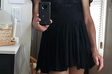 Picture of Miri standing in a doorway, wearing a very cute short black dress, smiling and holding a camera to take the picture of herself. She is hoping you like what you see.