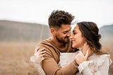 This image is of a couple sitting almost nose to nose in a field. The man is holding the woman’s face and they are looking at each other lovingly.