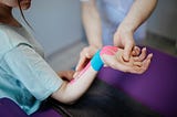 A person receiving kinesiology taping therapy on their wrist, with pink and blue tape applied, while a therapist adjusts their arm.