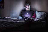 A blonde woman in a rumpled bed with her face illuminated by an open laptop