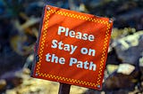 A picture of a red sign on a stake driven into the ground that says “Please stay on the path”.