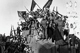 Black and white photograph of a crowd of revolutionaries, with raised rifles and fists, and flying CNT-FAI flags. Many are wearing red and black side caps (gorro milicianos).
