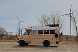 A small tan bus which has been converted into a tiny home, or skoolie, is parked in front of two large wind turbines.