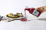 Disembodied hand pours beet red fermented drink into glass on white marble countertop