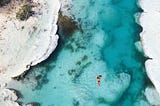 a drone from high above captures a person floating on their back in clear blue water between two large stromatolite formations