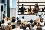 Police lineup being conducted by a robot in front of an audience of viewers. The lineup includes an aggressive male soldier, a female teacher, a large bear, and a skunk.