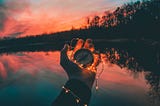 A compass surrounded by fairy lights and held by someone whose arm and palm can be seen, while behind it is a lake and a beautiful sunset.