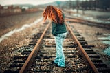 rear view of girl with long red hair wearing jeans and long sleeve jacket standing in middle of railroad track